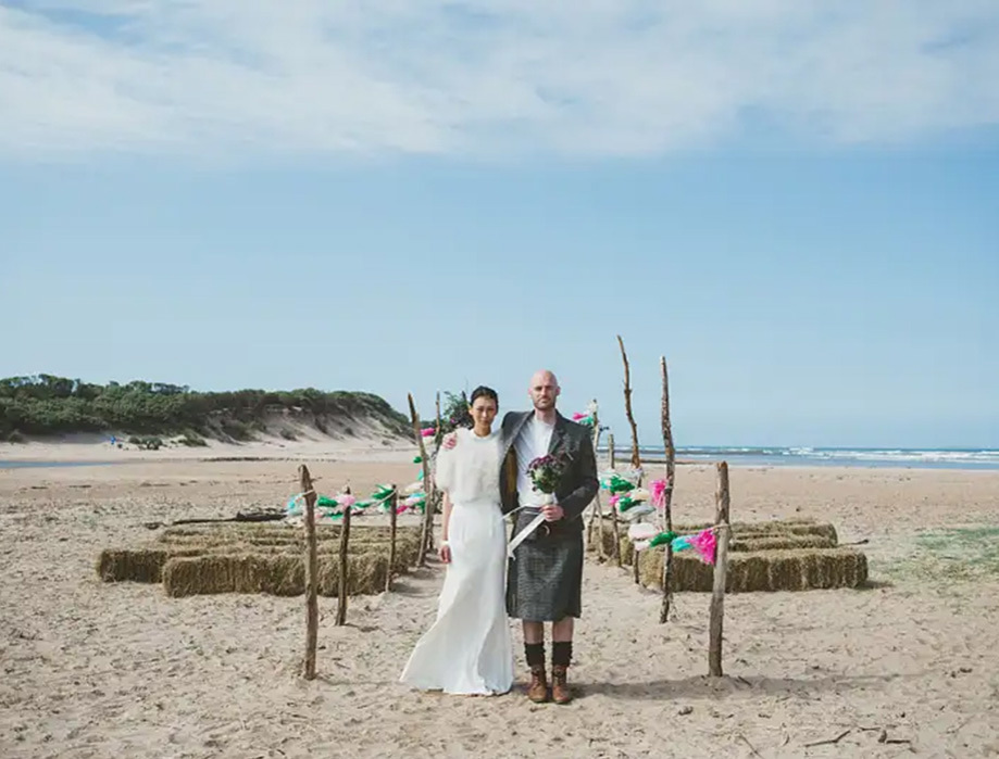 Bride and groom pose in front of their ceremony set up with hay bale seats on Tyninghame Beach
